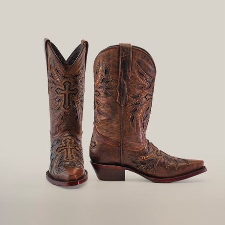 The Texas Cruz Tabaco boots exude style with intricate embroidery and cross motifs, crafted from premium leather. These womens mid-shaft boots feature a snip toe, angled heels, and decorative stitching. One boot stands upright while the other leans to the side against a plain white background.