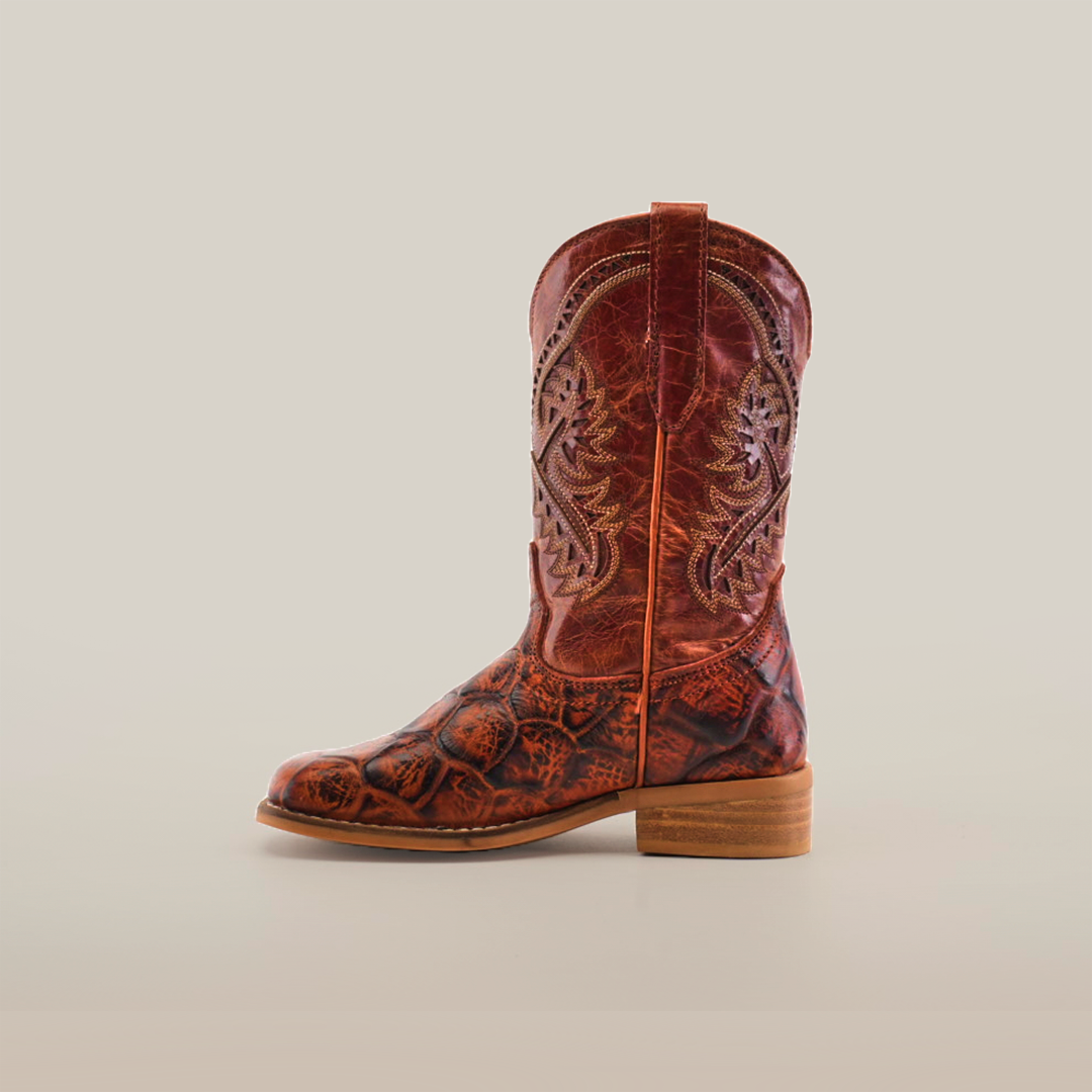 The Pirarucu Bass Print Cogñac Square Toe boot is a single brown cowboy boot showcasing ornate stitching and a textured Pirarucu bass print on the lower half. It features premium leather, a wooden heel, square toe, and pull tab, all set against a plain white background.