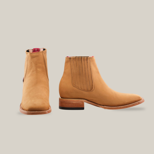 The Prime Suede Honey Square Toe boots, tan leather ankle style with a cowboy flair, are shown on a white background. One boot faces forward, the other displays its block heel and elastic side panels. Red back tabs add a pop of color to these fashionable essentials.