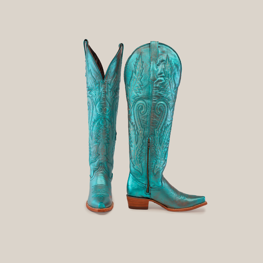 A pair of Brittany Rustic Ceramic Tall Shaft Snip Toe Boots in turquoise features intricate swirling embroidery, brown wooden heels, and a genuine leather sole. One boot stands upright while the other tilts, showing off the side zipper and pointed toe design.