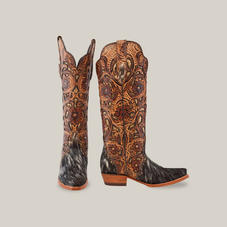 The Handtooled Cowhide Pinto boots are intricately designed womens cowboy boots featuring ornate tooled leather patterns and fur accents. These handcrafted boots boast a tall shaft, stacked heel, and blend brown hues with detailed craftsmanship against a light gray background.