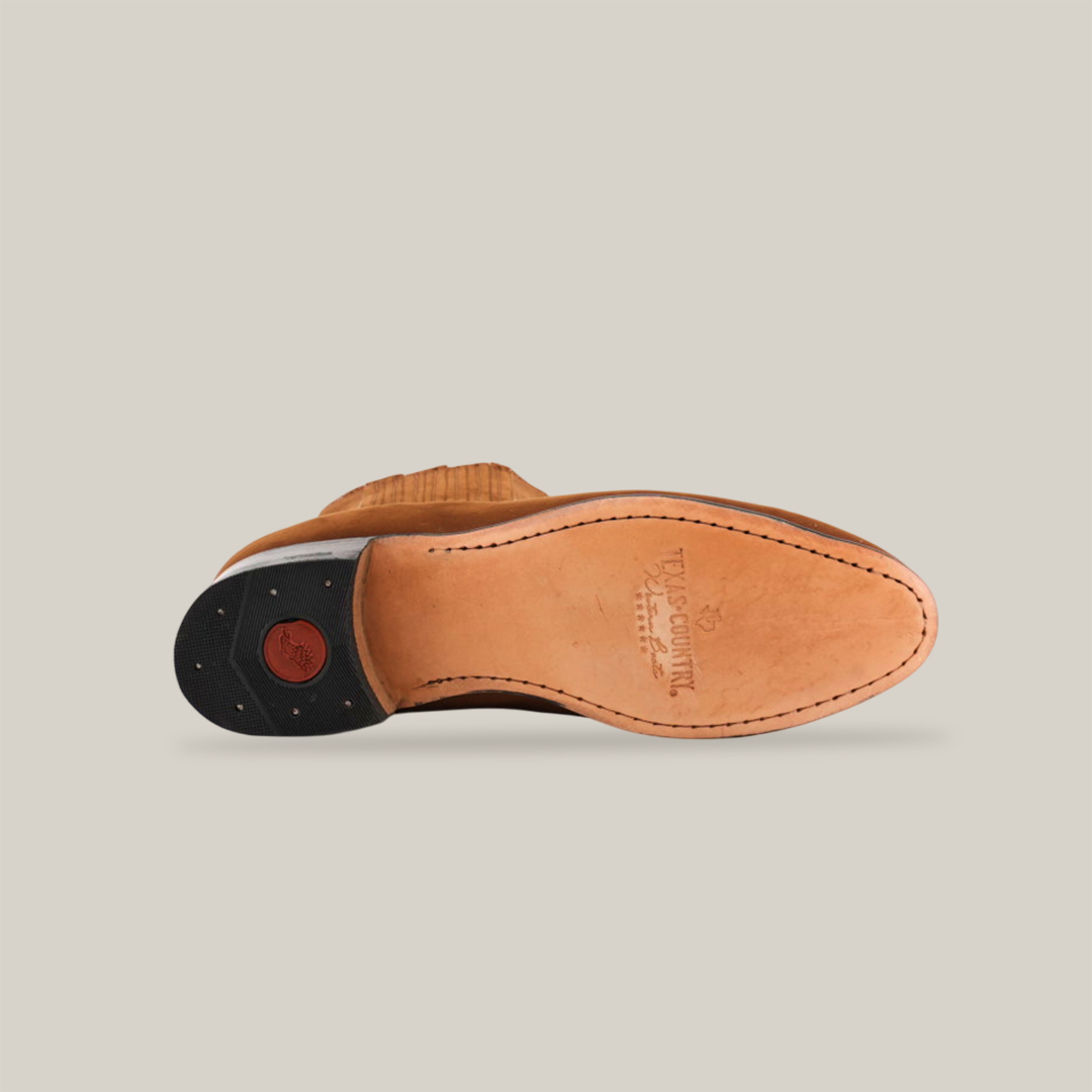 The bottom view of the Prime Suede Caramel Round Toe shoe reveals a stitched brown leather sole, a black heel, and a red circular detail reminiscent of Western style. The embossed logo at the center adds authenticity against a white background, highlighting its craftsmanship.