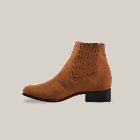 Introducing the Prime Suede Sand Round Toe, a brown marvel with red accents and elastic side panels on a crisp white background. This suede ankle boot features a low black heel, ideal for adding style and flair to any cowboy ensemble.