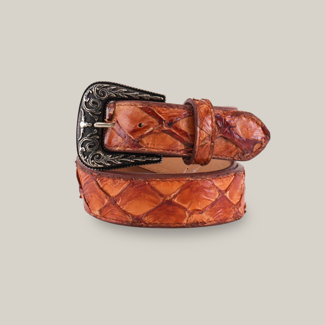 The Exotic Full Quilt Pirarucu Fish -Texas Buckle - Brandy Belt features a textured brown leather with an embossed pattern and a Texas-style silver buckle adorned with intricate floral engravings, demonstrating exceptional craftsmanship. It is artfully rolled up against a white background.