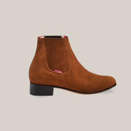 The Prime Suede Caramel Round Toe boot, featuring brown suede with a black heel and elastic panels, embodies Western charm. Its red and pink patterned lining adds flair, and its photographed against a white background to highlight its impeccable craftsmanship.