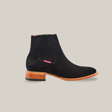 Explore the Prime Suede Black - Square Toe ankle boot, made from premium materials. This black suede boot includes a side elastic panel, brown leather sole, low heel with a dark rubber base, red stitching, and an embossed logo, offering a modern take on classic western style.