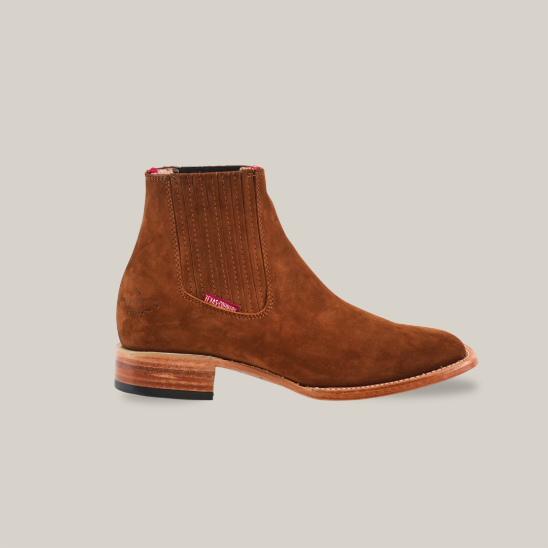 The Prime Suede Caramel Square Toe boots, with a Western touch and wooden soles, are featured on a white background. One boot stands upright and the other is turned to display intricate stitching, emphasizing this cowboy fashion staple.