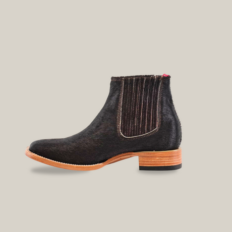 Side view of the Cowhide Hair Black Square Toe, a stylish black Chelsea boot in cowhide leather with a polished wooden sole and low heel. It features elastic side panels, a small red pull tab at the back, and is set against an off-white background.