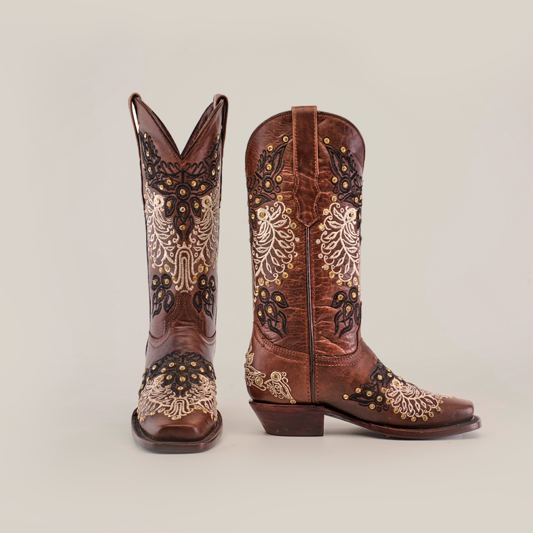 The Faenza Crystals Brown boots feature handcrafted rich brown premium leather, embellished with detailed white and yellow embroidery. Displayed against a plain background, one boot stands upright while the other sits angled, showcasing their mid-shaft and narrow square toe design.