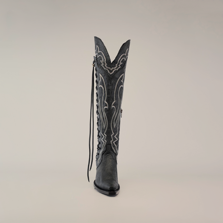 The Tania Matte Black boot is tall, handcrafted in matte black leather with intricate white stitching and a long side tassel against a plain light background, featuring a snip toe and decorative pattern