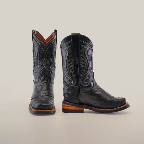 The Karoo Ostrich Half Cowhide Print Black Rodeo Toe boots feature a western aesthetic with intricate stitching and wooden soles. Displayed on a plain white background, one boot faces forward while the other is in profile, showcasing the detailed design.