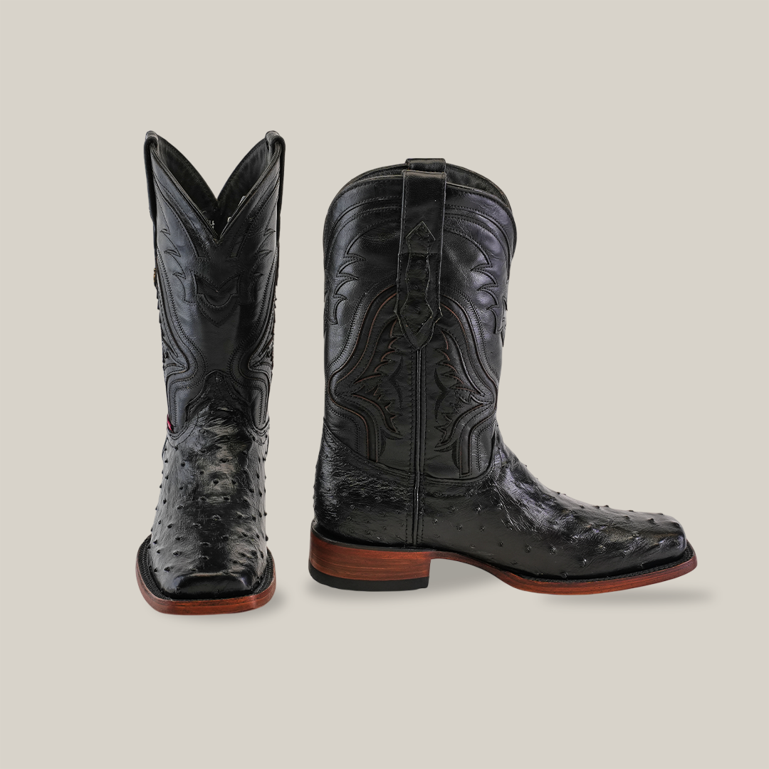 The Exotic Ostrich - Black - Rodeo Toe boots feature genuine ostrich leather with intricate stitching and textured patterns. One boot stands upright, the other angled, showcasing a wooden heel and pointed toe against a plain light background.