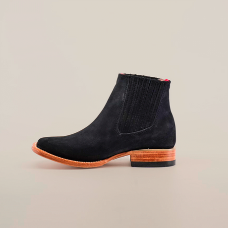 The Prime Suede Black Square Toe is a black suede ankle boot with a wooden sole, square toe, and low heel. It features elastic side panels and exudes elements of cowboy fashion, beautifully displayed against a plain white background to highlight its elegant minimalist design.