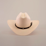 The 1000X Sinaloa Straw Hat features an elegant design with a wide brim, a black band adorned with silver conchos, and a plain white background.