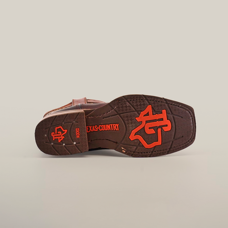 The image shows the sole of an Azkar Moka Slip Resistant Sole Rodeo Toe boot, adorned with an orange longhorn logo and TEXAS COUNTRY in bright text. The detailed tread provides slip resistance, and the brown cowboy boot rests on a clean white surface