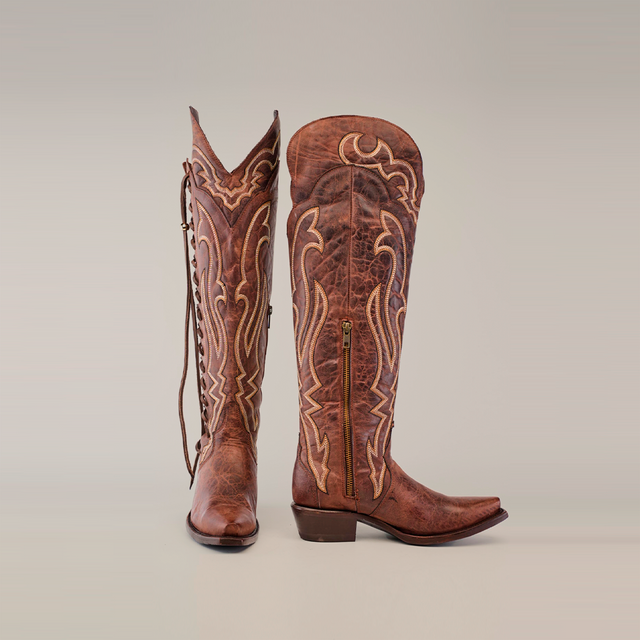 The Tania Brown boots, featuring a tall shaft and snip toe design, are crafted from luxurious materials with intricate white stitching and decorative patterns. One boot stands while the other is slightly tilted, both resting against a plain gray background.