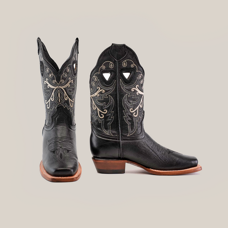 The Western Stitch Black Lustre boots feature ornate silver stitching, showcasing traditional craftsmanship. With narrow square toes and brown leather soles, one boot stands upright while the other is tilted to highlight its intricate side pattern.