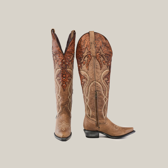 The Miriam Cappuccino Tall Shaft boots feature intricate embroidery and a tall decorative shaft, with one upright boot and the other leaning slightly, highlighting their handcrafted ornate patterns on the rich brown upper against a plain white background.