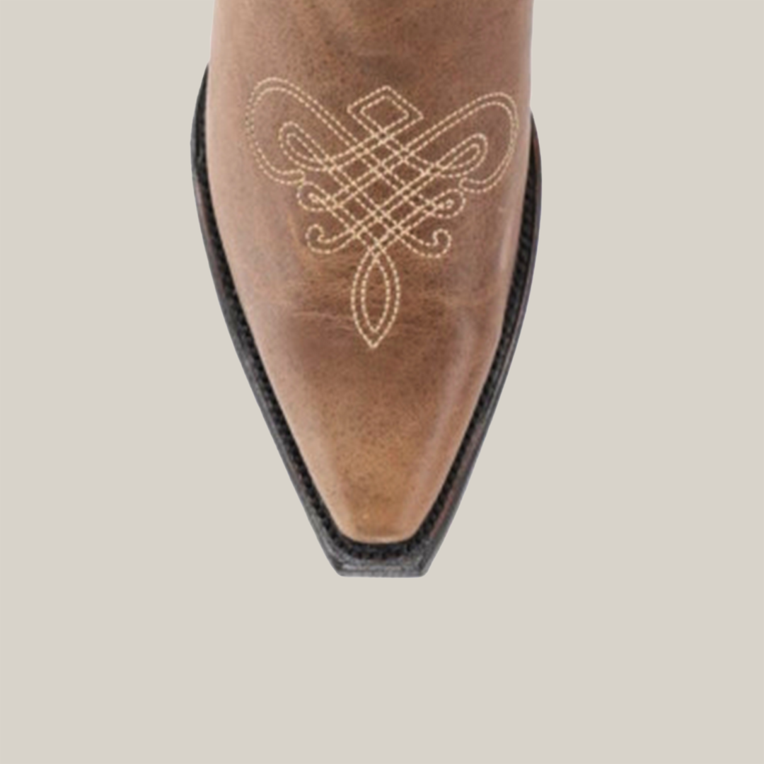 The Miriam Cappuccino boot features a snip toe with intricate white stitching that exemplifies artisanal craftsmanship on brown leather. Its black sole complements the detailed design, all beautifully showcased against a plain white backdrop.