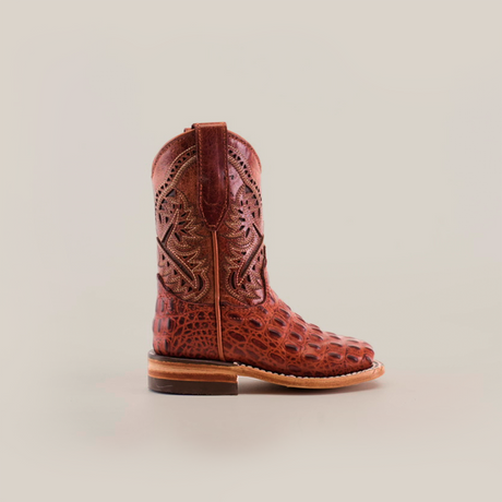 The Caiman Hornback Print Cogñac Square Toe boot, featuring a textured reptile-like pattern and intricate stitching, graces a wooden surface. Its premium brown leather finish stands out against the plain light background, highlighting its quality and craftsmanship.