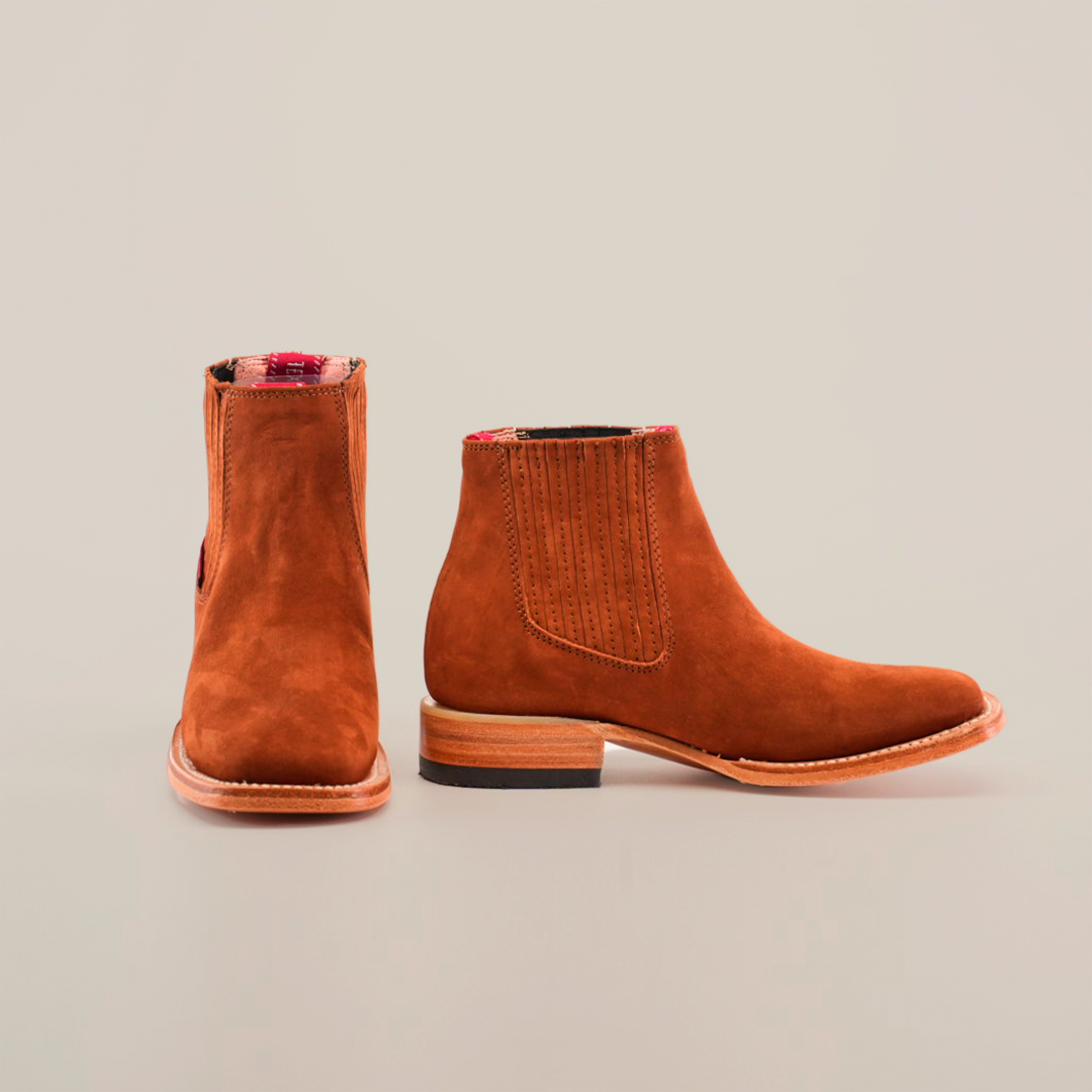 The Prime Suede Brick Square Toe boots feature brown suede with flat heels, red plaid lining peeking from the top, and elastic side panels for easy slip-on. One boot faces forward while the other displays its side profile in a classic cowboy style.