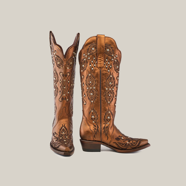 The Rania Copper boots, with their tall shaft and snip toe design, are handcrafted in brown leather, featuring intricate embroidery and silver studs. Set against a plain white background, one boot stands upright while the other is angled slightly forward.