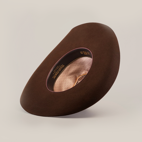 The 6X Brazil Brown felt cowboy hat is angled to reveal its inside brim and decorative lining with a patterned label. It casts a subtle shadow, highlighting its depth and texture against a light background, showcasing its Western heritage.