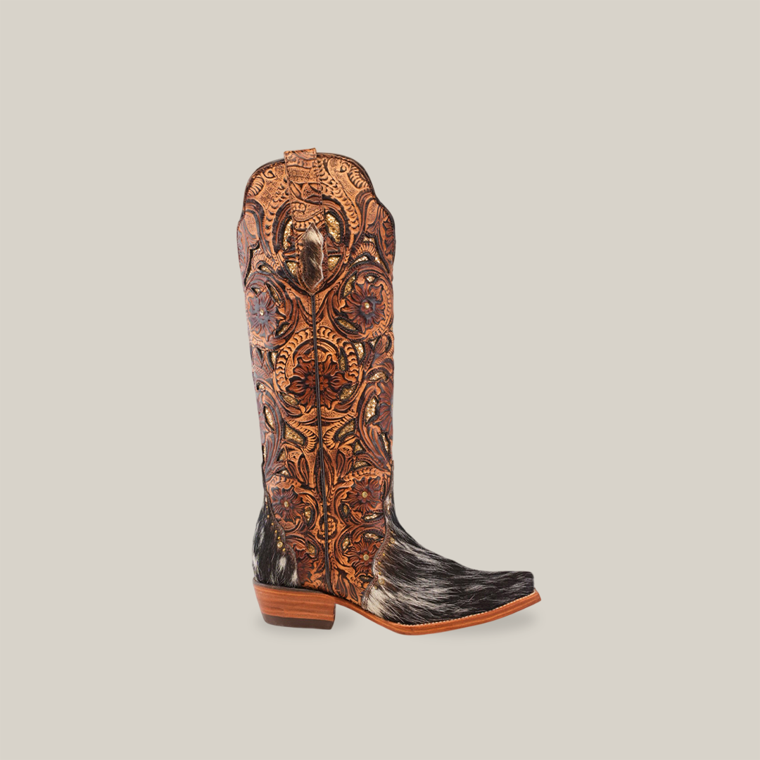 The Handtooled Cowhide Pinto - Tall Shaft - Snip Toe boots feature intricate brown leather tooling and dark speckled hair-on-hide details, set against a light gray background. Handcrafted with precision, these boots exude rustic elegance.