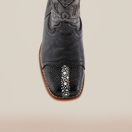 The Stingray Half Cowhide Print Black Rodeo Toe boot, made from cowhide leather, showcases western charm with its intricate toe pattern and white stitching. It features a brown sole and detailed shaft design, displayed from above against a plain backdrop.