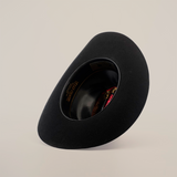 The X 2 Carnales Black, a classic wide-brimmed felt hat, rests upside down on a plain white background. Its inner lining is slightly visible, showing hints of gold text and red fabric.