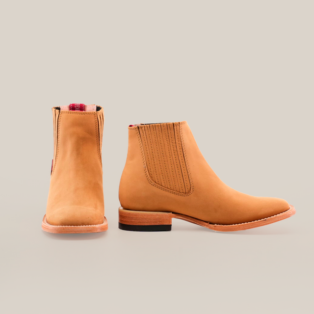 The Prime Suede Honey Square Toe boots are tan, ankle-length, and feature elastic side panels, a square toe, and light-colored soles. Displayed on a white background, they evoke cowboy style with one boot facing forward and the other to the side.