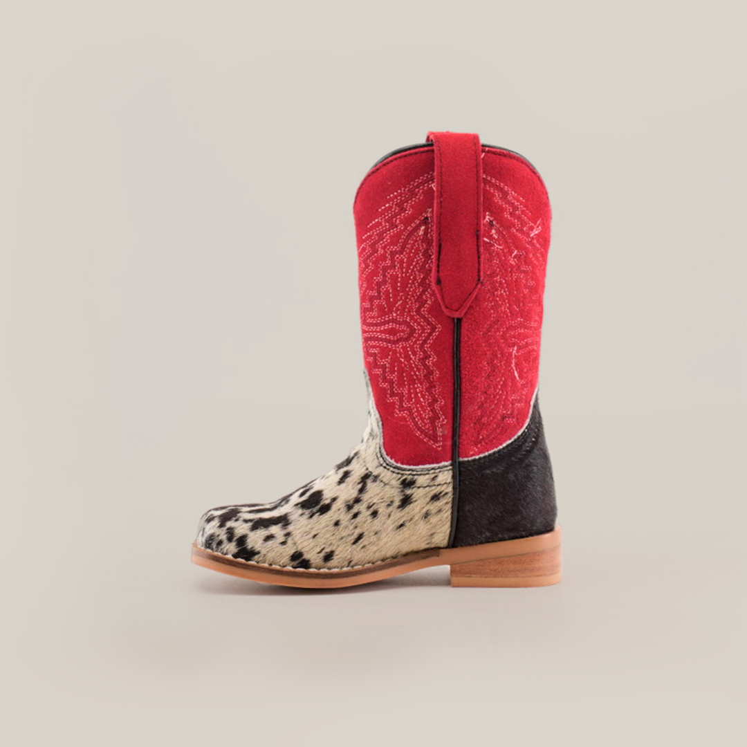 The Cowhide Hair Pinto Square Toe childrens cowboy boot features a western style, with a red shaft adorned with embroidered star patterns and a spotted black and white cowhide hair pinto shoe, complete with a light brown sole. The boot is shown from the side against a white background.