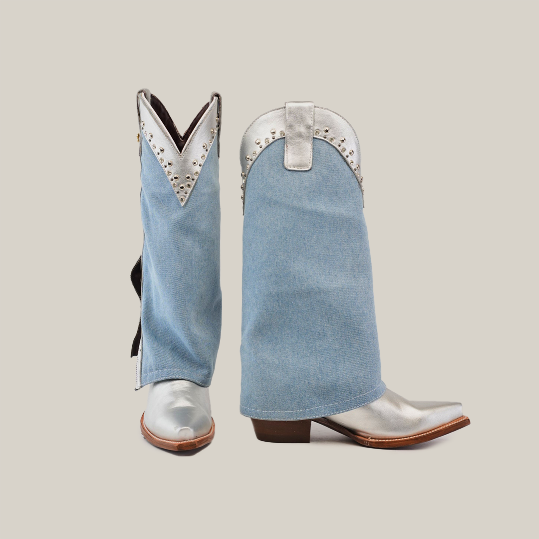 The Bella Denim Silver boots, with a tall shaft and metallic snip toes, stand side by side. Adorned with decorative metal studs, they blend rustic and modern fashion against a plain background.