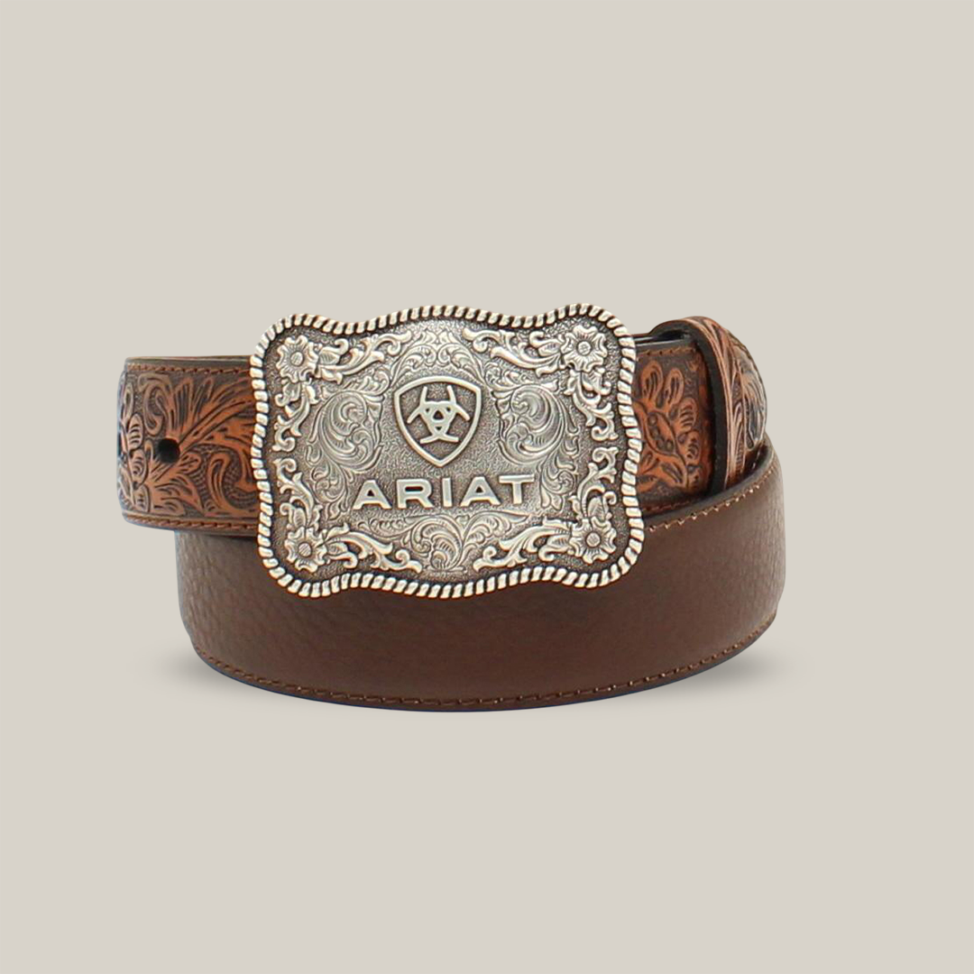 The Ariat Boys Belt Flower Tooled Brown - A1301002 is a brown leather belt with floral embossing, featuring a large rectangular silver buckle engraved with patterns and the ARIAT logo, making it a standout Western-inspired accessory.