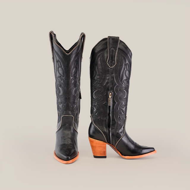 The Linda Studs Tall Shaft Black - J Toe boots from the Platinum Collection are luxury handcrafted cowboy boots with intricate patterns, decorative studs, a tall shaft, side zipper, and stacked wooden heels. One boot is upright while the other is angled to highlight its artistry.