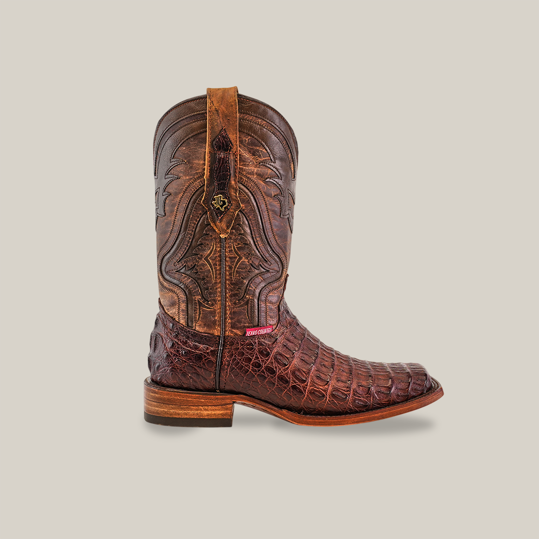 The Exotic Caiman Hornback - Brown - Rodeo Toe boot is crafted with caiman leather, featuring intricate stitching, crocodile texture on a plain background, detailed hornback accents, and a small red label near the sole for unique charm.