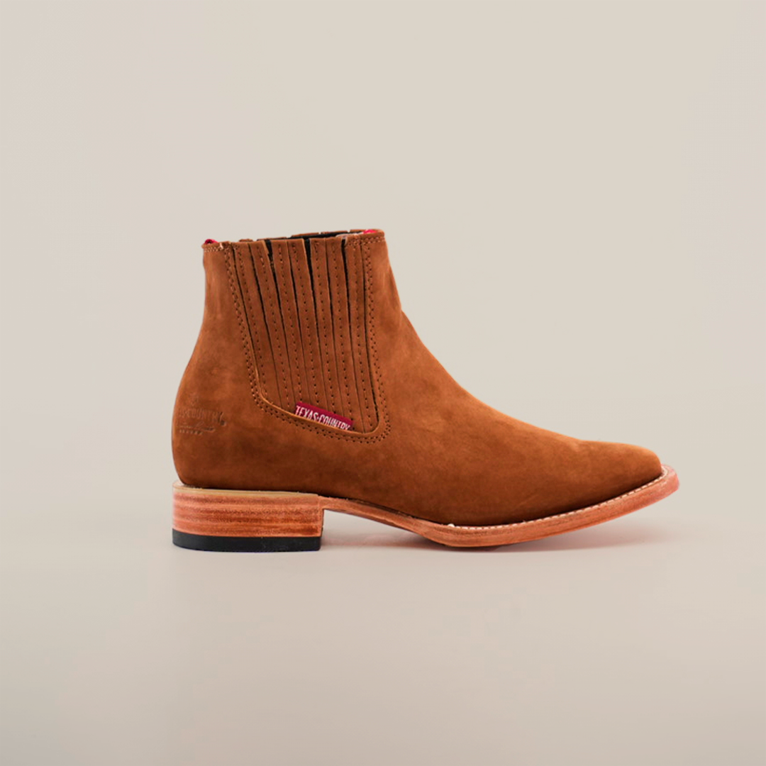 The Prime Suede Caramel Square Toe boot is brown with elastic side panels, a modest heel, and a red detail near the top. It has a square toe and is showcased on a cowboy scene-inspired white background.