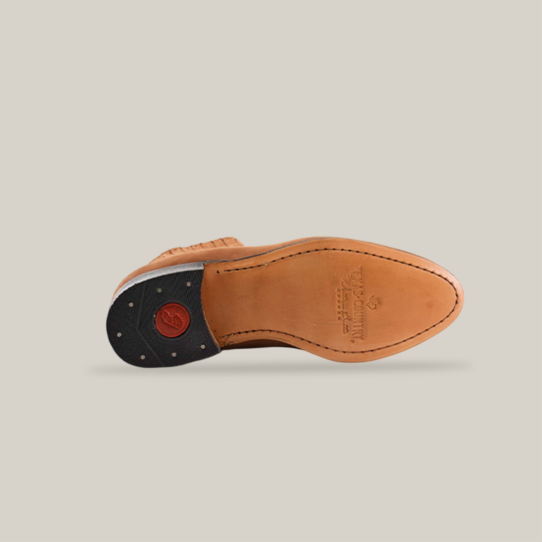 A Prime Suede Sand Round Toe shoe in tan leather rests on its side, evoking a classic cowboy style. The sole features detailed edge stitching and a black heel with a red emblem, set against a plain, light background.