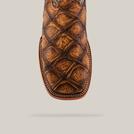 The Big Bass Pirarucu Print Rustic Honey cowboy boot, handcrafted in square-toed brown leather with premium cowhide, boasts a textured crisscross pattern and detailed stitching along the edge against a plain white background.