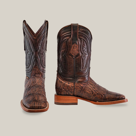 A pair of Gator Print Rustic Brown cowboy boots features intricate stitching and a rustic leather look. The left boot stands upright, while the square-toe right boot leans slightly, highlighting its heel. Both boots have a textured finish with flat wooden soles.