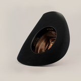 The 6X Bull Black hat, featuring an asymmetrical wide brim and a patterned brown interior, is set upright on a white background. Its unique shape exudes an artistic, modern vibe and captures the luxury essence of western wear.