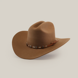 The 6X Sinaloa Fawn, a brown cowboy hat with a wide brim and decorated band with round metal accents and a side knot, embodies rich Western heritage. Crafted from premium sheeps wool, it is showcased on a plain white background.