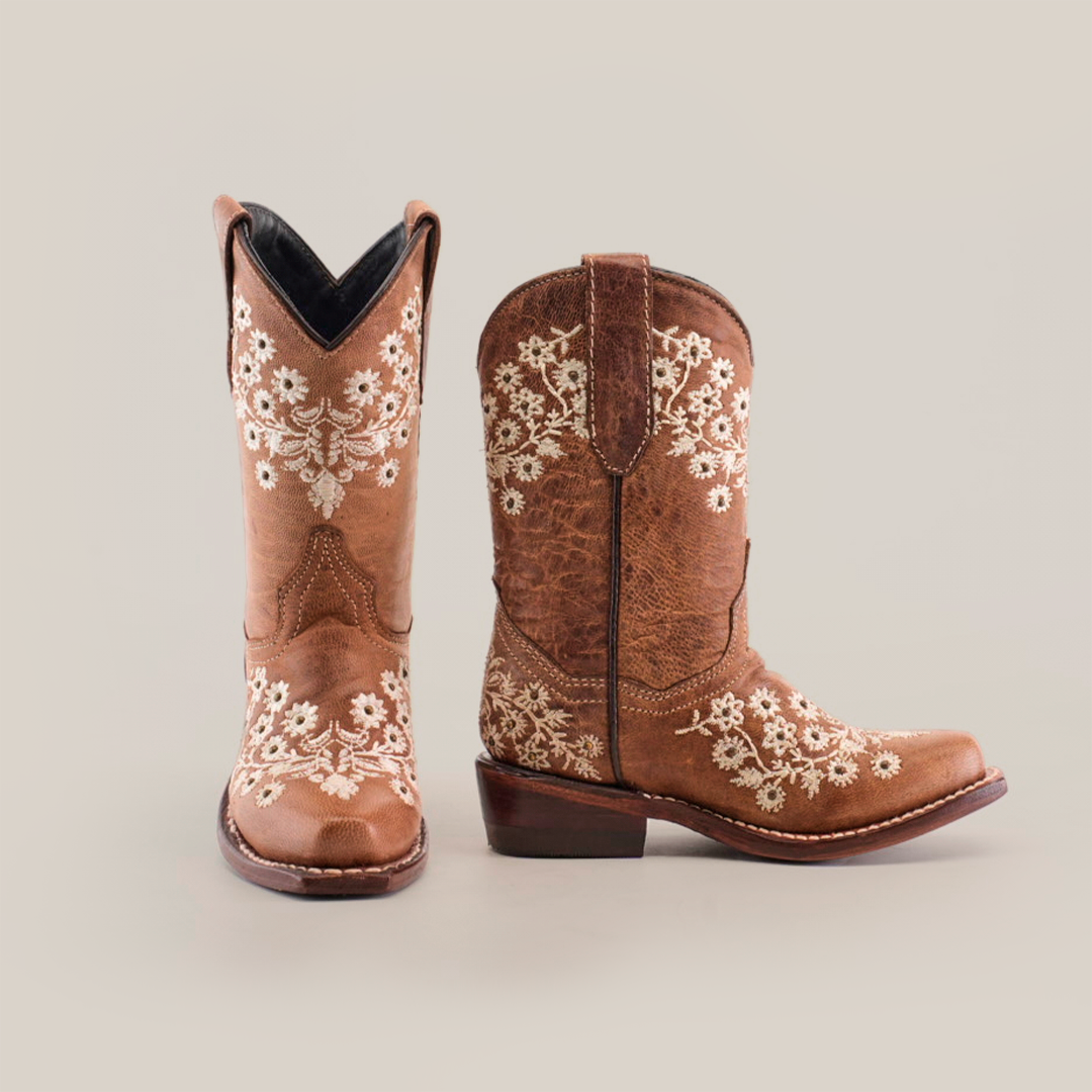 The Abril Studs Orix Snip Toe boots are brown leather cowboy boots with intricate white floral embroidery on the sides and front, featuring a stylish snip toe and stacked heel, showcased against a plain white background.