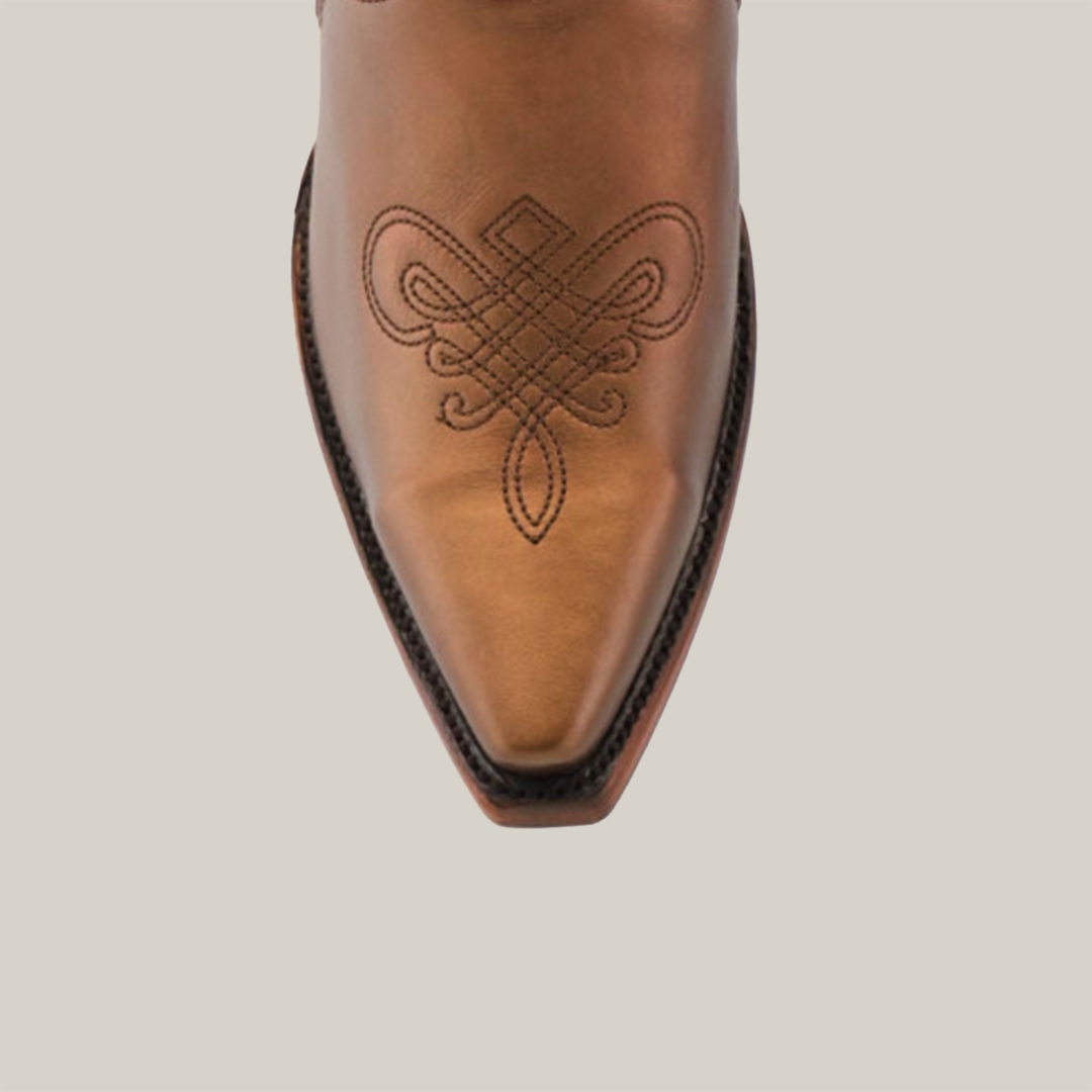 A close-up of a brown leather cowboy boot, similar to Nihan Copper - Tall Shaft - Snip Toe, displays intricate embroidered swirls. The handcrafted quality is evident in the pointed snip toe and dark stitching along the edges against a plain backdrop.