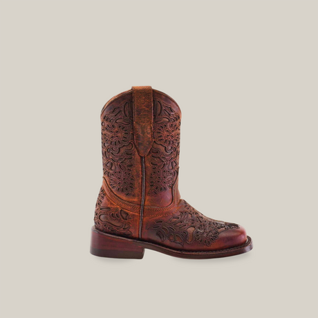 The Yennis Brick Square Toe, a girls cowboy boot, showcases intricate floral embossing. Crafted from premium brown leather, it stands upright against a white background with a low heel and convenient side pull straps for easy wear.