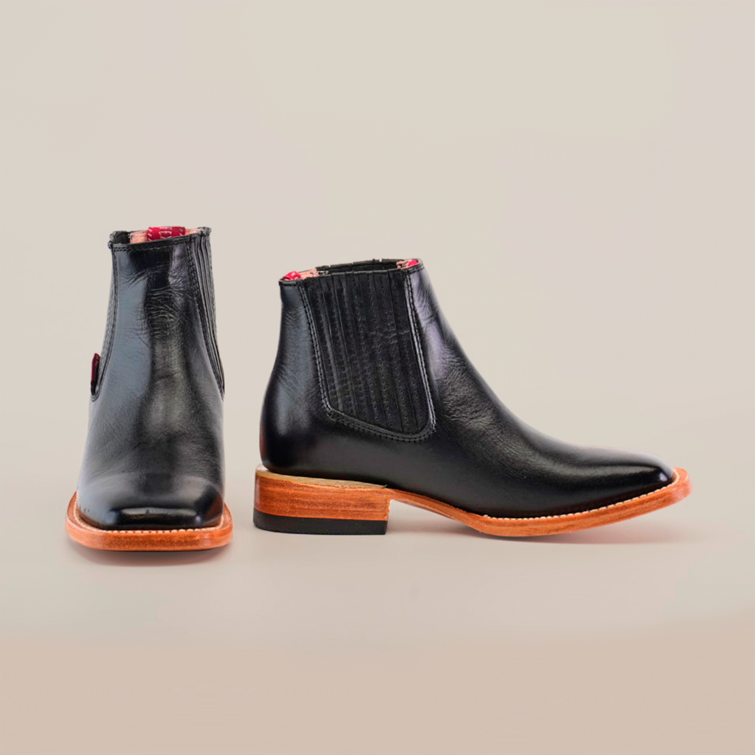 The Napa Black Square Toe boots feature wooden soles, with one boot showcasing the front and sole and the other at an angle to highlight its elastic side panel. The inside has a chic red pattern, making it ideal for any fashion-forward wardrobe.