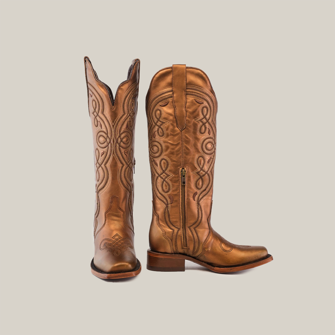 The Eden Copper Tall Shaft boots, with a narrow square toe, showcase intricate brown leather embroidery and a side zipper. One boot stands upright while the other is angled side-on, highlighting their luxury craftsmanship and block heel design.