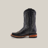 The Barcelona Black Lustre Rodeo Toe boot, made from premium cowhide leather, boasts intricate stitching with a brown stacked heel. This black boot has a wide square toe and pull tabs, all highlighted against a white background.