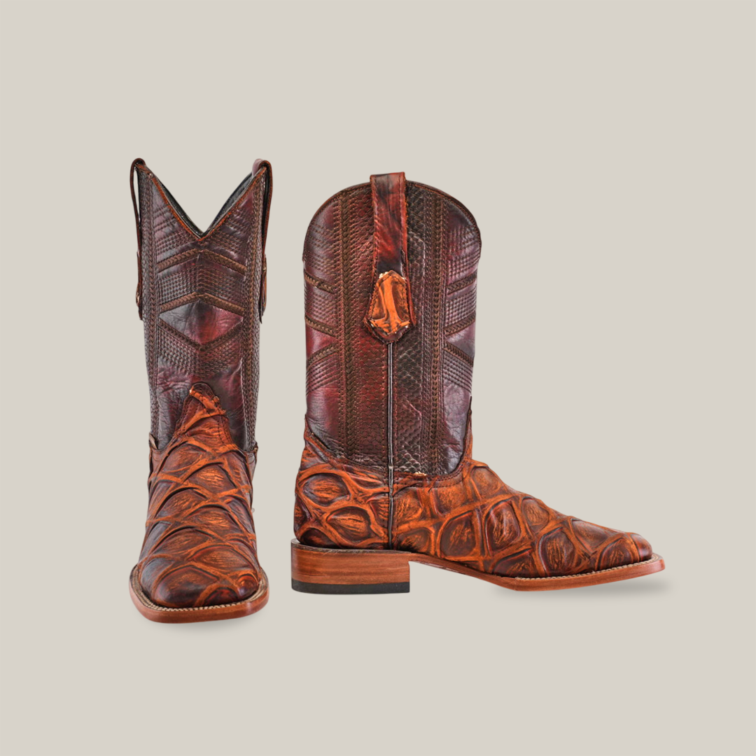 The Big Bass Pirarucu Print Rustic Cogñac square toe boots, crafted from premium brown cowhide, showcase intricate patterns with distinct leather designs. They feature traditional high shafts and pull straps, set against a plain white background.