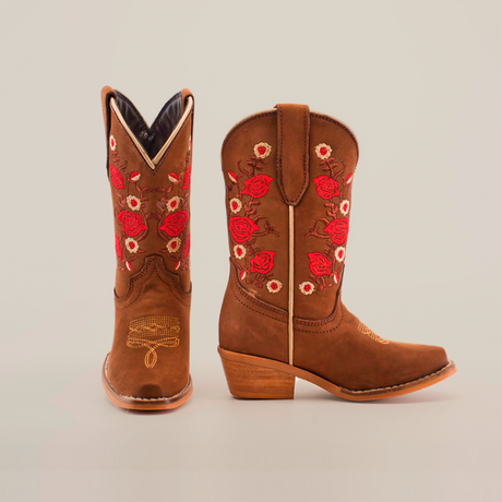 The Mia Copper Suede Snip Toe boots are brown, featuring red rose embroidery and snake designs. Handcrafted from premium leather, they have wooden heels and intricate stitching. One boot stands upright; the other lies flat to highlight detailing.